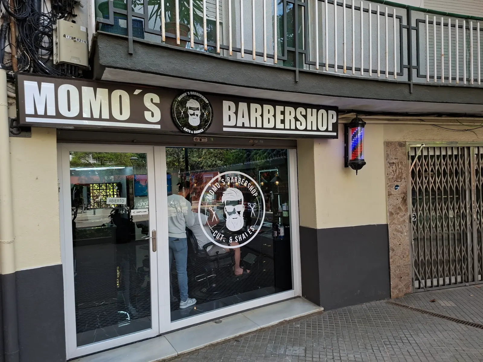 Fachada de Momo's Barbershop: rótulo negro con el logo, puerta de cristal y el poste de barbero